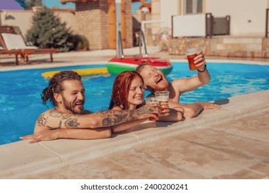 Group of cheerful young friends having fun at the swimming pool, leaning on the edge while making a toast with glasses of beer, relaxing while on a summer vacation - Powered by Shutterstock