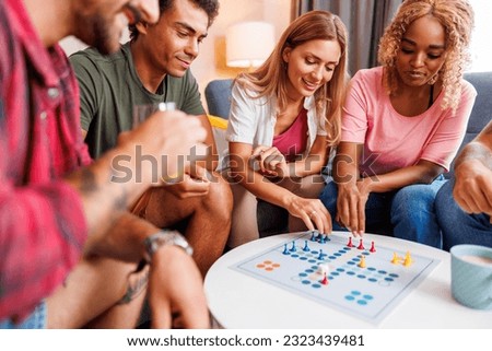 Group of cheerful young friends having fun playing ludo board game while spending leisure time together at home