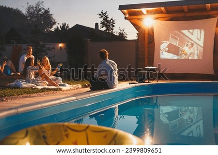 Group of cheerful young friends having fun eating popcorn, drinking cocktails and watching a movie in a home backyard poolside open air cinema