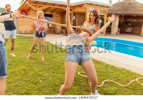 Group of cheerful young\
friends having fun at summertime outdoor party by the swimming\
pool, participating in limbo dance contest, passing below the rope\
while dancing