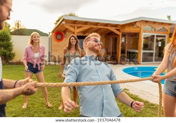 Group of cheerful young\
friends having fun at summertime outdoor party by the swimming\
pool, participating in limbo dance contest, passing below the rope\
while dancing