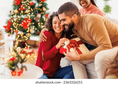 Group of cheerful young friends spending Christmas together at home, having fun exchanging and opening presents - Powered by Shutterstock