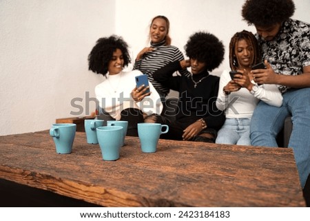 Group of cheerful young men and women laughing while looking at smartphone and watching funny videos on social media, at home. Multiethnic people sitting on sofa having fun together.