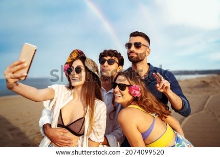 Group of cheerful young people taking a selfie on the beach with a rainbow in the background - summer vibes with friends having fun in the summer