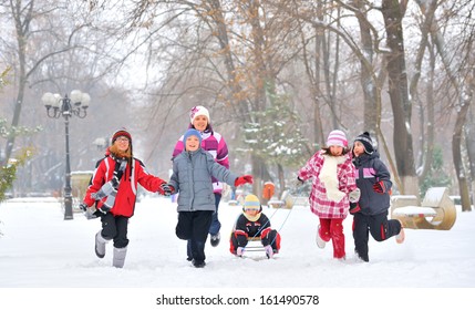 Group Of Children And Adult Playing On Snow In Winter Time, Young Girl Pulling Sister Through Snow On Sled