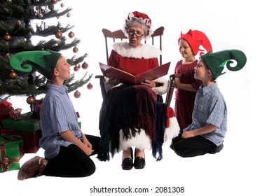 A Group Of Children Gathered Around Mrs. Santa Claus Sitting In Her Rocking Chair And Reading Them A Story.