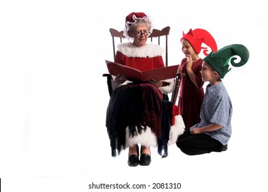 A Group Of Children Gathered Around Mrs. Santa Claus Sitting In Her Rocking Chair And Reading Them A Story.