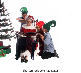 A Group Of Children Gathered Around Mrs. Santa Claus Sitting In Her Rocking Chair And Reading Them A Story.