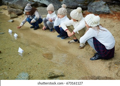 Group Of Children Playing Outdoors: Siting On Lake Shore By Clear Water And Sending Paper Ships To Float On Warm Autumn Day Dressed In Similar Knit Clothes