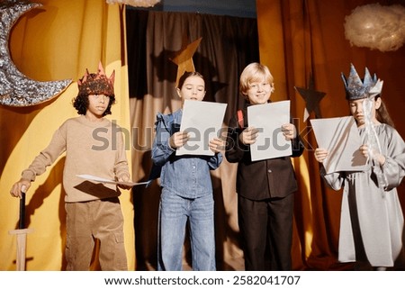 Group of children reading scripts during school play performance, holding papers, and wearing unique costumes with star and moon decorations positioned in background