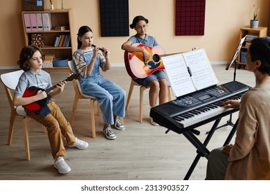 Group of children sitting on chairs and playing musical instruments with teacher playing piano during lesson in music class - Powered by Shutterstock