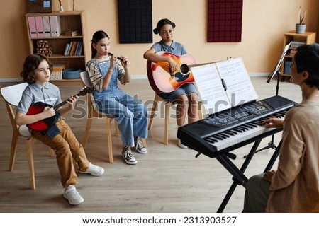 Group of children sitting on chairs and playing musical instruments with teacher playing piano during lesson in music class