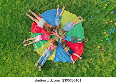 Group of children with teachers holding hands together on rainbow playground parachute in park, top view. Summer camp activity - Powered by Shutterstock