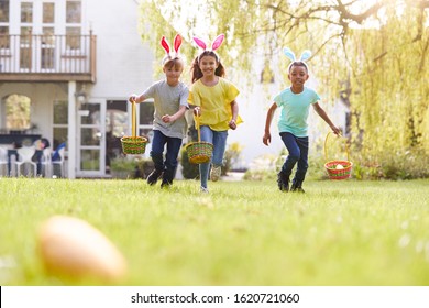 Group Of Children Wearing Bunny Ears Running On Easter Egg Hunt In Garden