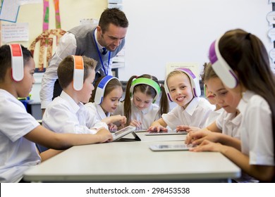 Group Of Children Wearing Colorful Wireless Headsets While Working On Digital Tablets, The Teacher Can Be Seen Supervising The Students In The Classroom