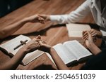 Group of Christians sit together and pray around a wooden table with blurred open Bible pages in their homeroom. Prayer for brothers, faith, hope, and seek the blessings of God.
