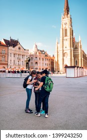 Group Of Classmates Standing In Town Square And Looking At Smart Phone On School Excursion