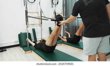 Group of clients performing Pilates exercises on Cadillac reformer machine for improving core strength and leg engagement. Healthy client posing lower and lift pose while trainer assisting. Habituate. - Powered by Shutterstock