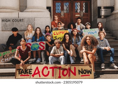 Group of climate change activists looking at the camera while sitting outside a high court with posters and banners. Multicultural young people protesting against global warming and pollution. - Powered by Shutterstock