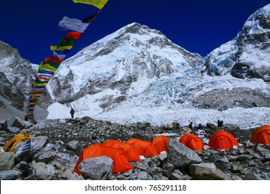 Group of climbers ' bright color tents on the Khumbu glacier in area of Everest base camp with colorful prayer flags and himalaya mountain range in background during a clear blue sky day