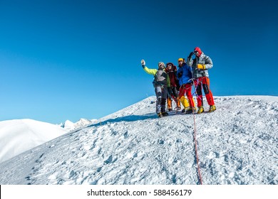 Group Of Climbers In Protective Insulated Outwear Alpine Gear And Ropes Rising Hands Enjoying The Victory Against Severe High Altitude Mountain