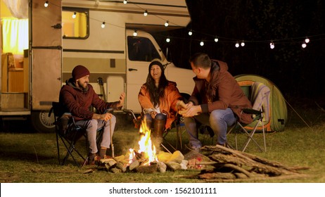 Group of close friends laughing together around camp fire. Retro camper van. Light bulbs in the background.