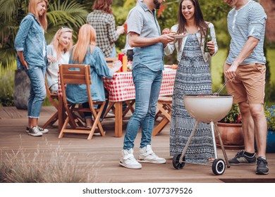 Group Of Colleagues Having A Summer Party On A Garden Patio, Sitting By A Table And Standing By A Grill