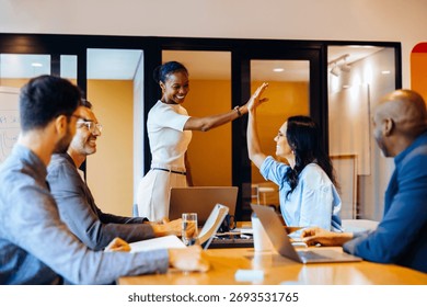 Group of colleagues seated at a table while discussing projects, cheering with high fives. Joyful expressions reflect successful communication in a dynamic and productive office environment.