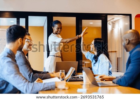 Group of colleagues seated at a table while discussing projects, cheering with high fives. Joyful expressions reflect successful communication in a dynamic and productive office environment.