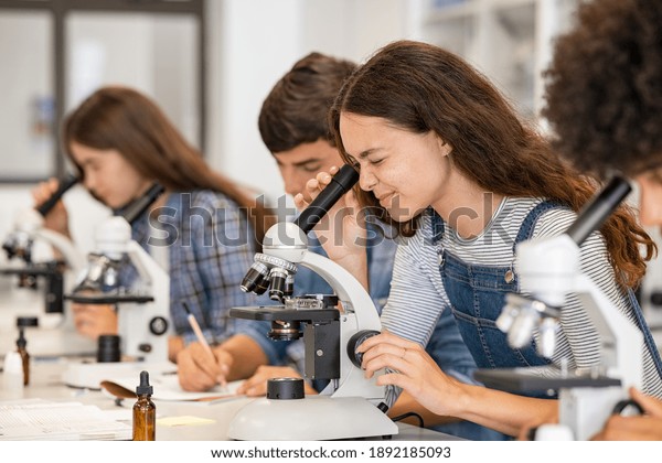 Group of college students performing experiment\
using microscope in science lab. University focused student looking\
through microscope in biology class. High school girl examine\
samples during lecture