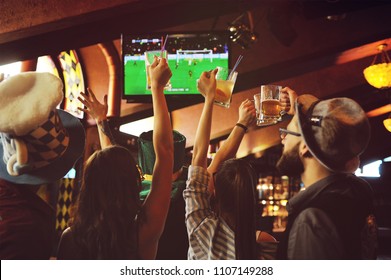 Group Or Company Of Friends - Young Guys And Girls Holding Glasses Of Beer, Watching Football, Laughing And Smiling At The Bar During The Oktoberfest Festival