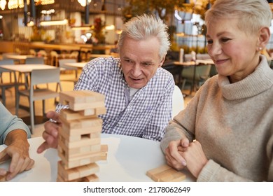 Group Of Concentrated Senior Friends Sitting At Table In Nursing Home Leisure Room And Playing Jenga