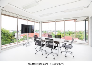 Group Of Conference Table And Chairs In White And Wide Office With Television Screen Without Anyone In Room, Green Tree Outside