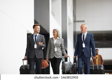 Group Of Confident Company Managers In Suits Walking With Luggage In Airport And Discussing Business Trip