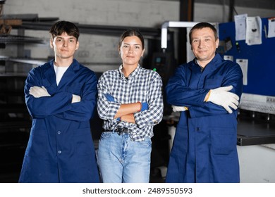 Group of confident smiling workers, young woman and two men, posing together in industrial metalworking workshop, showcasing teamwork and professionalism in manufacturing environment.. - Powered by Shutterstock