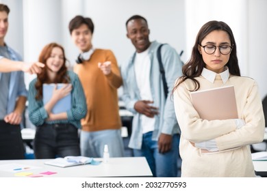 Group Conflict. Sad Latin Woman In Eyeglasses Standing Away Alone, Suffering Humiliation And Public Disgrace, Crowd Of Bad People Bullying Pointing Fingers At Victim, Laughing. Discrimination, Gossip