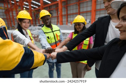 Group of construction workers clad in hard hats and reflective vests, are locked in a cordial fist bump, signaling joint progress. Machinery and steel beams compose the factory backdrop.