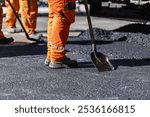 A group of construction workers is engaged in paving a road, using shovels to spread fresh asphalt. The sun shines bright on this bustling urban worksite.