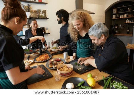 group of cooks, friends or colleagues cut ingredients cook together and prepare dinner at a wooden table in a kitchen or restaurant