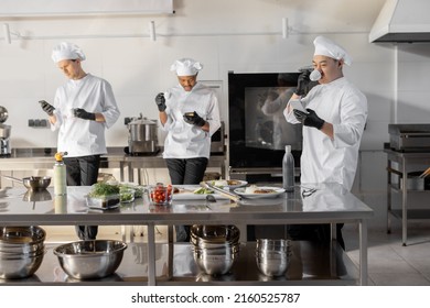 Group Of Cooks Using Phones And Drinking Coffee During A Break In The Restaurant Kitchen. Professional Staff Resting After Work In Kitchen. Multiracial Team Of Cooks Indoors