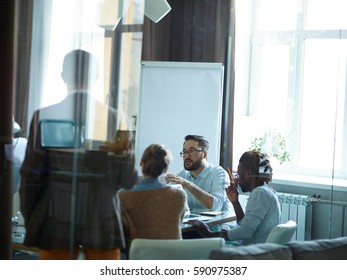 Group Of Creative Young People, Afro-American Man Among Them, Sitting Round Meeting Table In Modern Conference Room Listening Intently To Asian Speaker, Shot From Behind Glass Wall