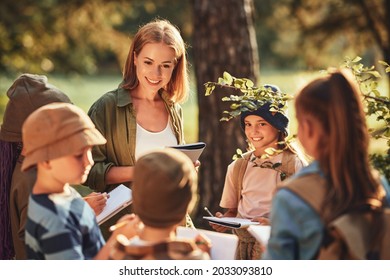 Group of curious school children with notebooks listening to their young female teacher while learning about nature together, looking at green leaf during ecology lesson in autumn forest on sunny day - Powered by Shutterstock