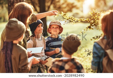 Group of curious school children with notebooks listening to their young female teacher while learning about nature together, looking at green leaf during ecology lesson in autumn forest on sunny day