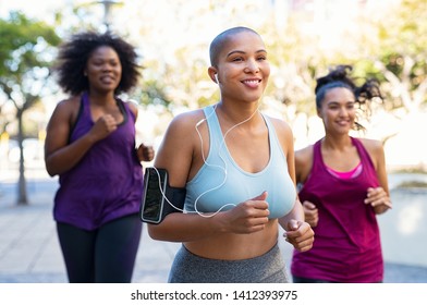 Group Of Curvy Women Jogging On Track In Park. Girls Friends Running Together Outdoor. Portrait Of Young Bald Woman Jogging With Oversize Friends While Listening To Music On Mobile Phone.