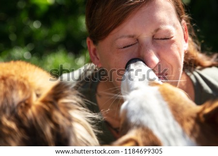 a group of cute funny dogs are licking the face of a beautiful young woman, outdoors with green plants in the background