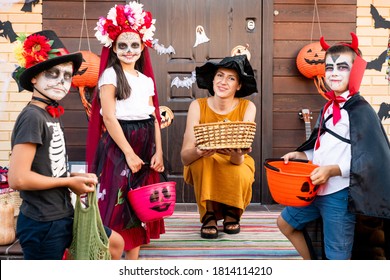 Group Of Cute Kids In Halloween Costumes Surrounding Young Smiling Woman With Basket Of Treats Sitting On Squats Against The Door