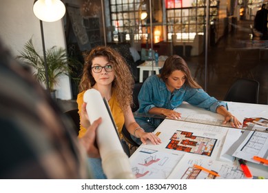 Group Of Designers Working On The Project. Female Designer With Curly Hair Reaching For The Blueprint.