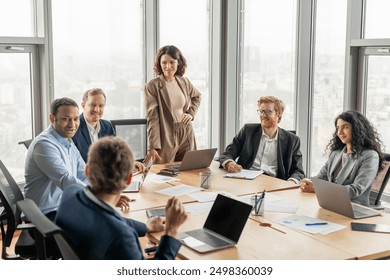 A group of diverse business professionals are seated around a large conference table in a modern office space. One woman stands at the head of the table, while the others focus on the discussion - Powered by Shutterstock