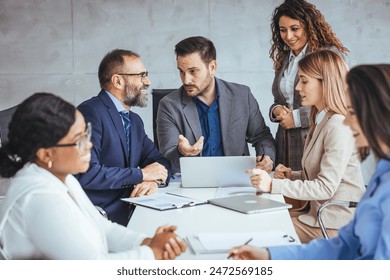A group of diverse business professionals in smart attire discuss strategies around a laptop in a modern office setting, actively exchanging ideas and notes.