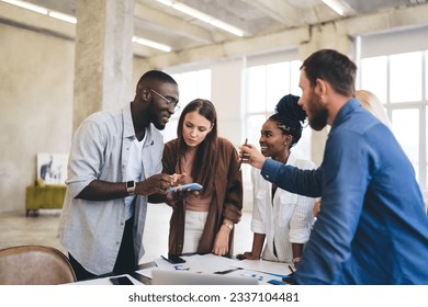 Group of diverse businesspeople standing at table with papers and demonstrating cellphones to each other while working on business startup in modern office together - Powered by Shutterstock